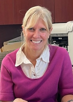Tracy Hudson sitting at a desk smiling wearing a pink sweater